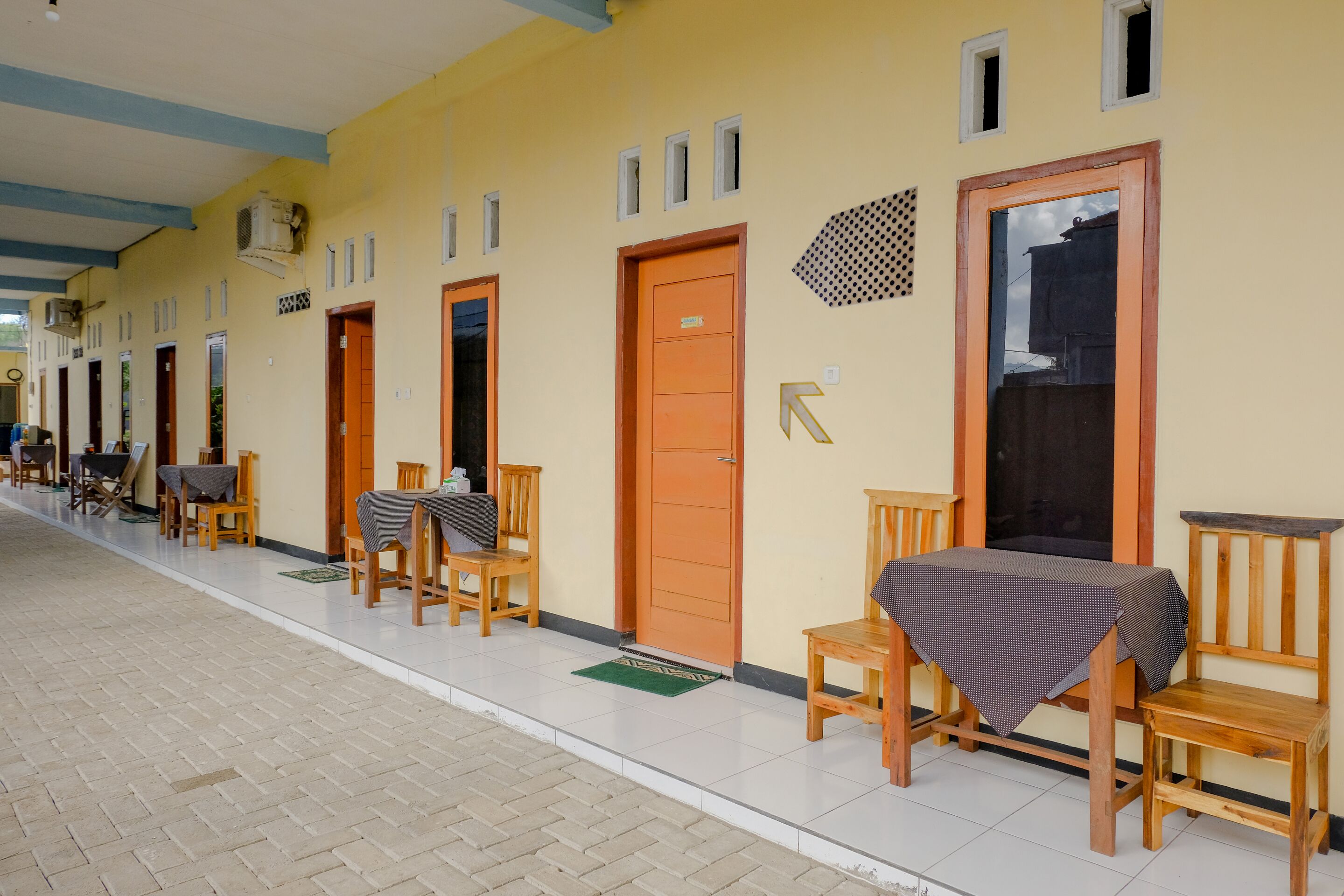 A corridor with wooden chairs and tables, doors, and a tiled floor.