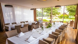 A dining area with multiple tables set for a meal, large windows offering a view of the outdoors, and wicker chairs with cushions. - Ahauser Land-&Golfhotel (Ahaus)