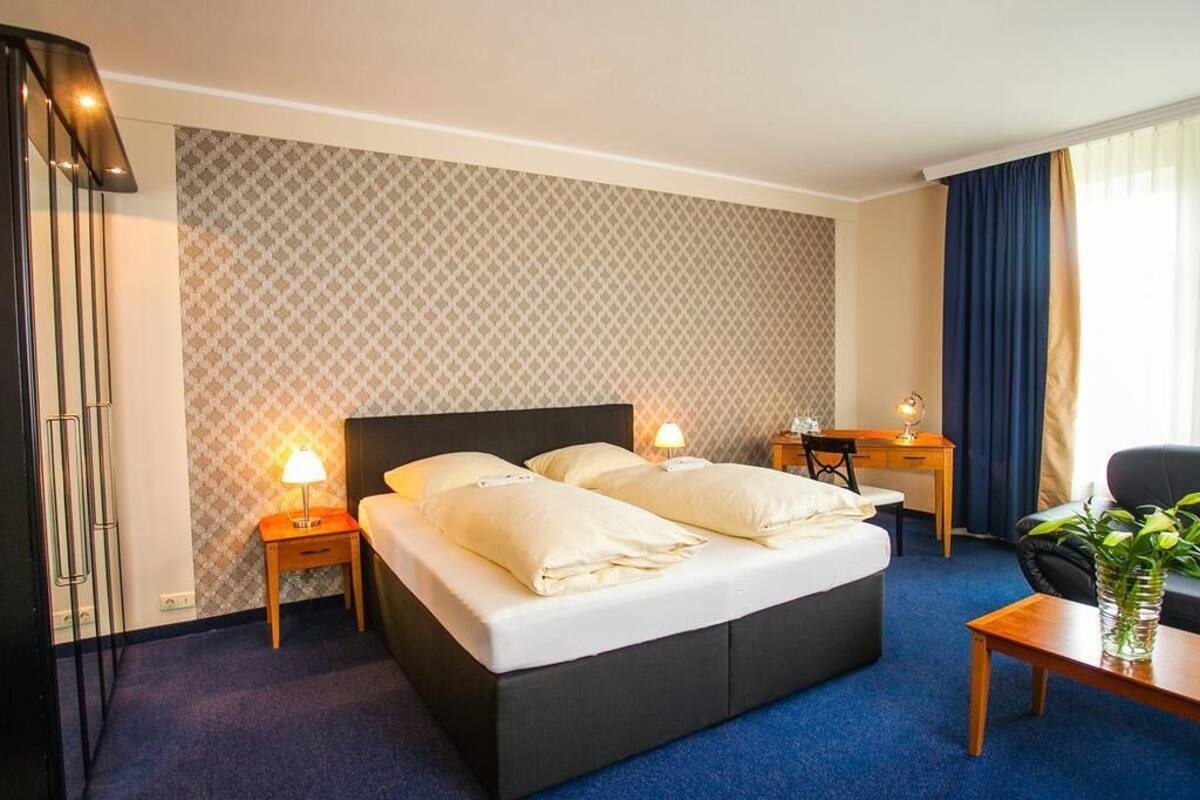 A hotel room with a large bed, two bedside tables with lamps, a desk, a chair, and a potted plant.