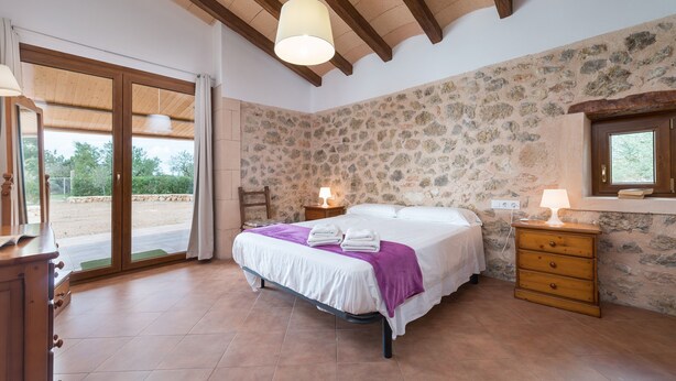 Une chambre avec un mur en pierre, un lit recouvert d’une courtepointe blanche et violette, une commode en bois et une grande fenêtre avec des rideaux.