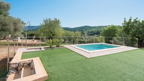 Une piscine rectangulaire bordée de pierres, entourée de gazon artificiel, avec en arrière-plan des arbres et des collines.