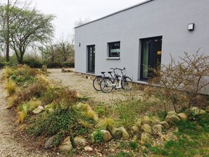 A modern building with a gravel driveway, two bicycles parked outside, and a natural landscape with rocks and shrubs. - Beltblick (Fehmarn)