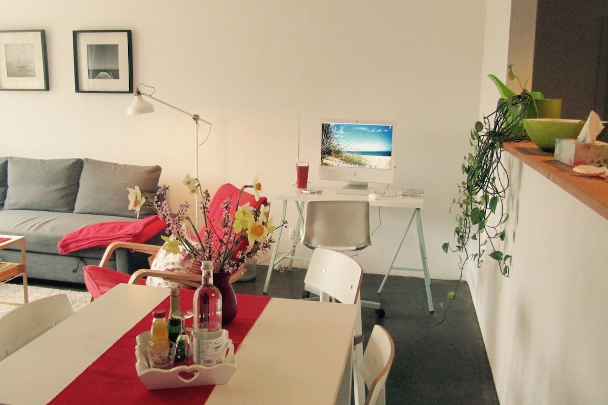 A living room with a sofa, a dining table with a vase of flowers, a desk with a computer, and a chair.