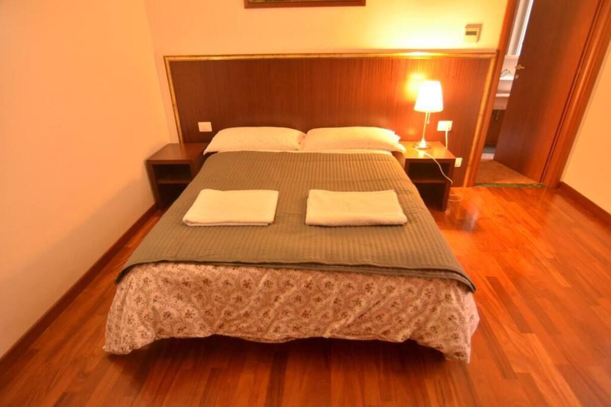 Cama bem arrumada com colcha floral e duas toalhas dobradas, abajur e cabeceira de madeira em quarto de hotel com piso de madeira.