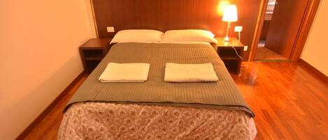 A neatly made bed with a floral bedspread and two folded towels, a bedside lamp, and a wooden headboard in a hotel room with wooden flooring.
