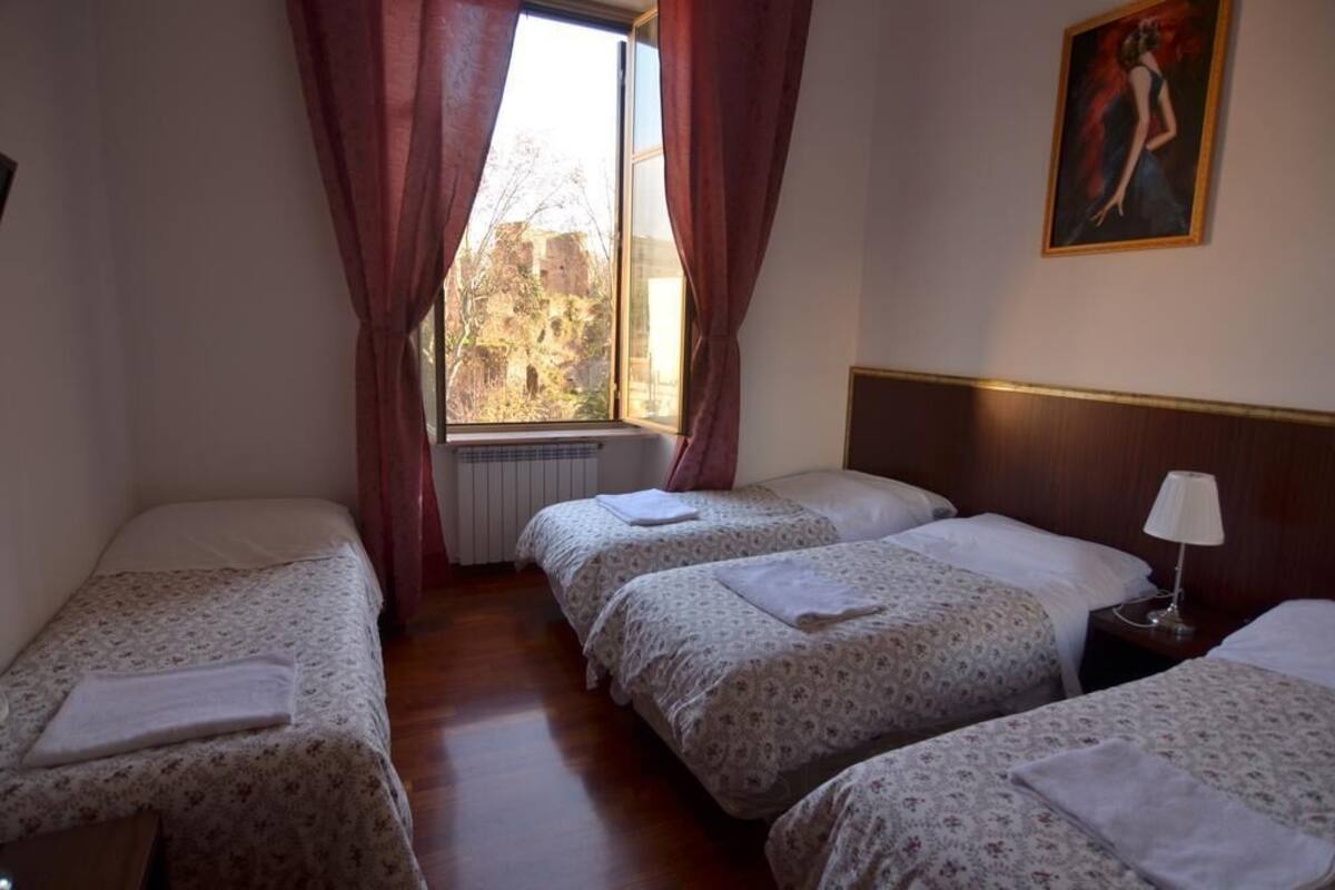 Quarto de hotel com três camas de solteiro, uma janela com cortinas, um radiador e um quadro na parede.