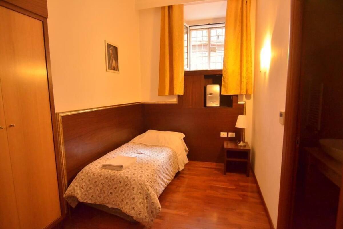 Quarto pequeno e acolhedor com uma cama de solteiro, mesa de cabeceira com abajur e janela com cortinas.