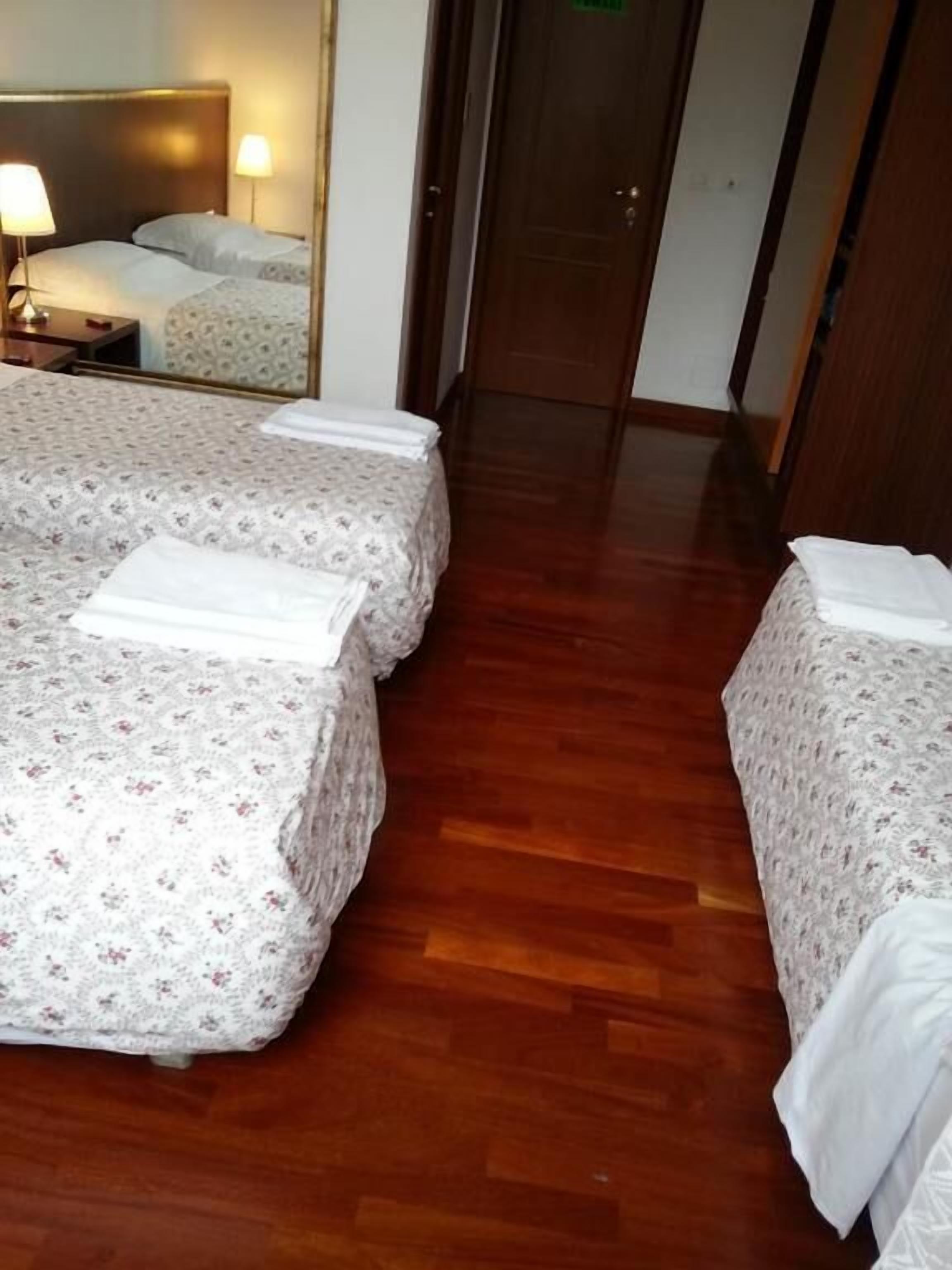 Een hotelkamer met twee bedden, elk met een bloemenprint sprei en witte lakens. Er ligt een houten vloer en er is een deur naar een andere kamer.