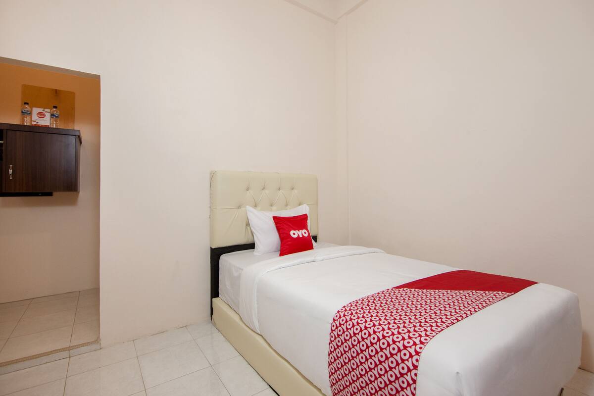 A hotel room with a bed, a headboard, a red and white patterned bedspread, and a small shelf with water bottles and a sign.