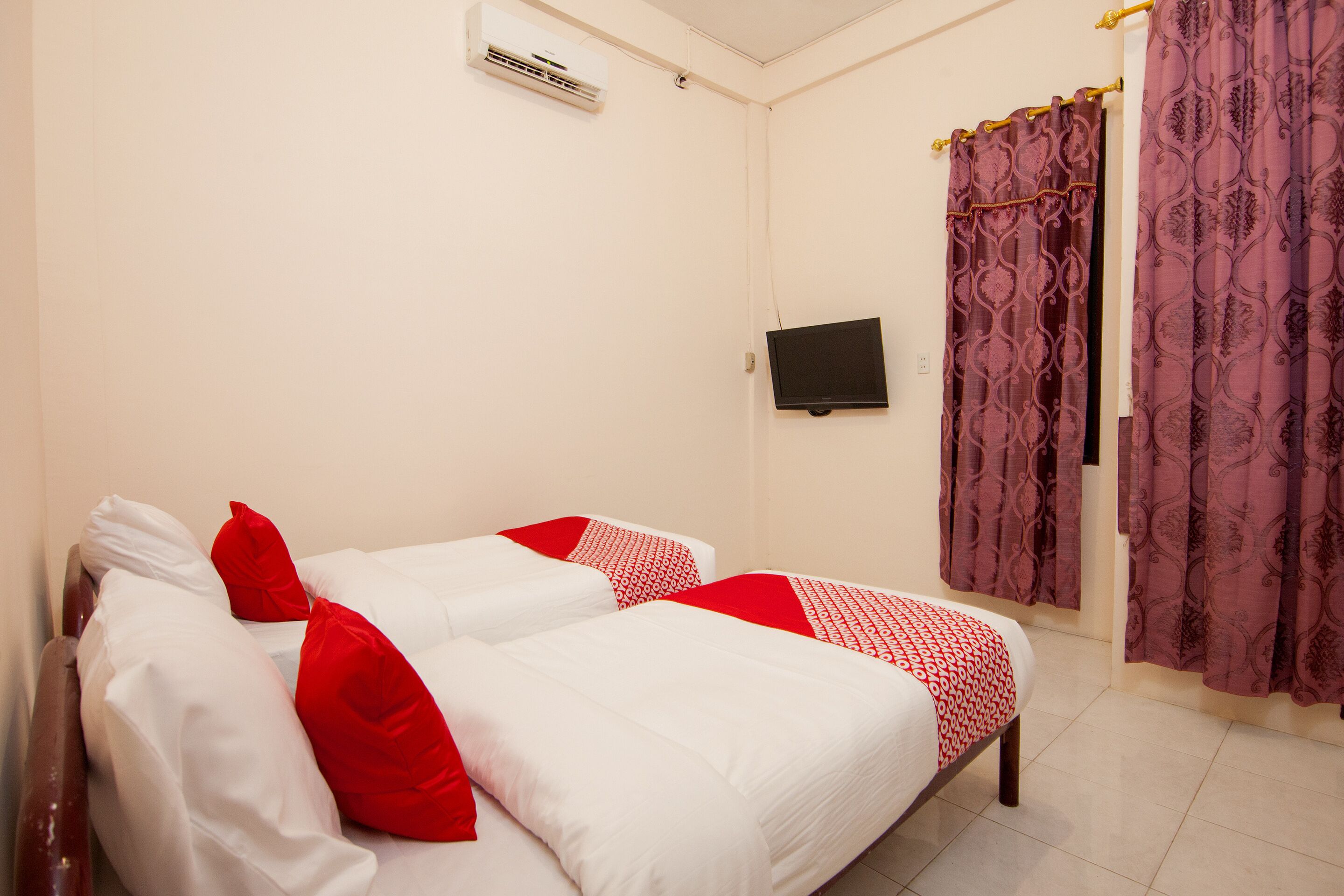 A room with two beds, each with white and red bedding, a flat-screen TV mounted on the wall, and purple patterned curtains.