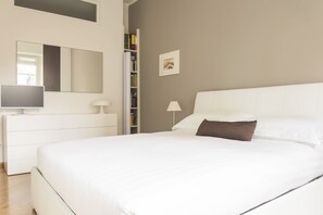 A bedroom with a large bed, a bedside table with a lamp, a bookshelf, and a mirror on the wall.