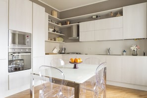 A modern kitchen with white cabinetry, a built-in oven, and a dining table with clear chairs.