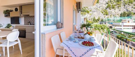 A balcony with a dining table set for breakfast, a kitchen with modern appliances, and a scenic view of a hillside.