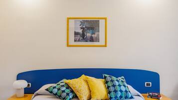 Ein Bett mit blauer Kopfteile, mehreren Kissen, einem Nachttisch mit Lampe und einem gerahmten Bild an der Wand.