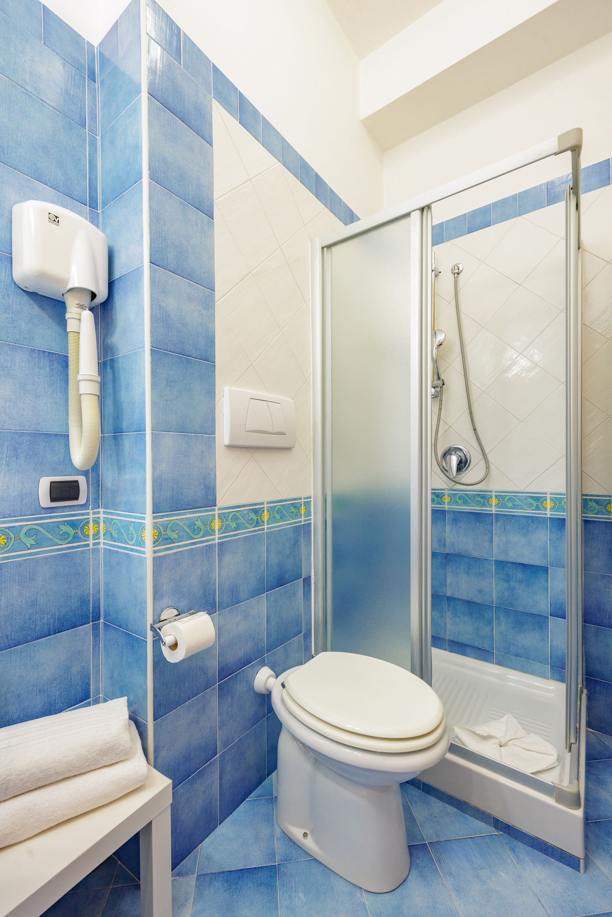 A bathroom with blue tiles, a toilet, a shower with a glass door, and a hand dryer.
