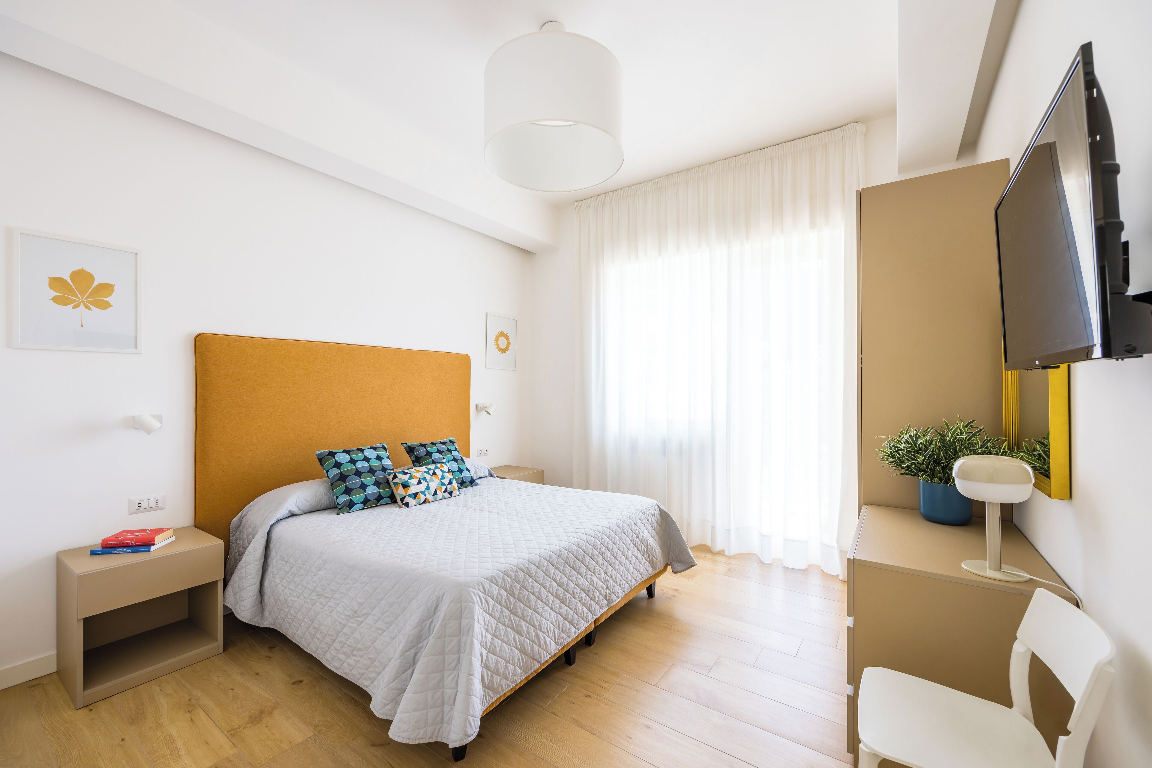 A modern bedroom with a bed, bedside tables, a desk, a chair, a television, and a window with curtains.