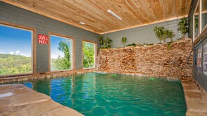 An indoor swimming pool with a stone wall, wooden ceiling, and large windows offering a view of the outdoors. - Ridge Top Retreat: Mtn Views, Indoor Pool, Theater, Games, Hot Tub, Wifi (Sevierville)