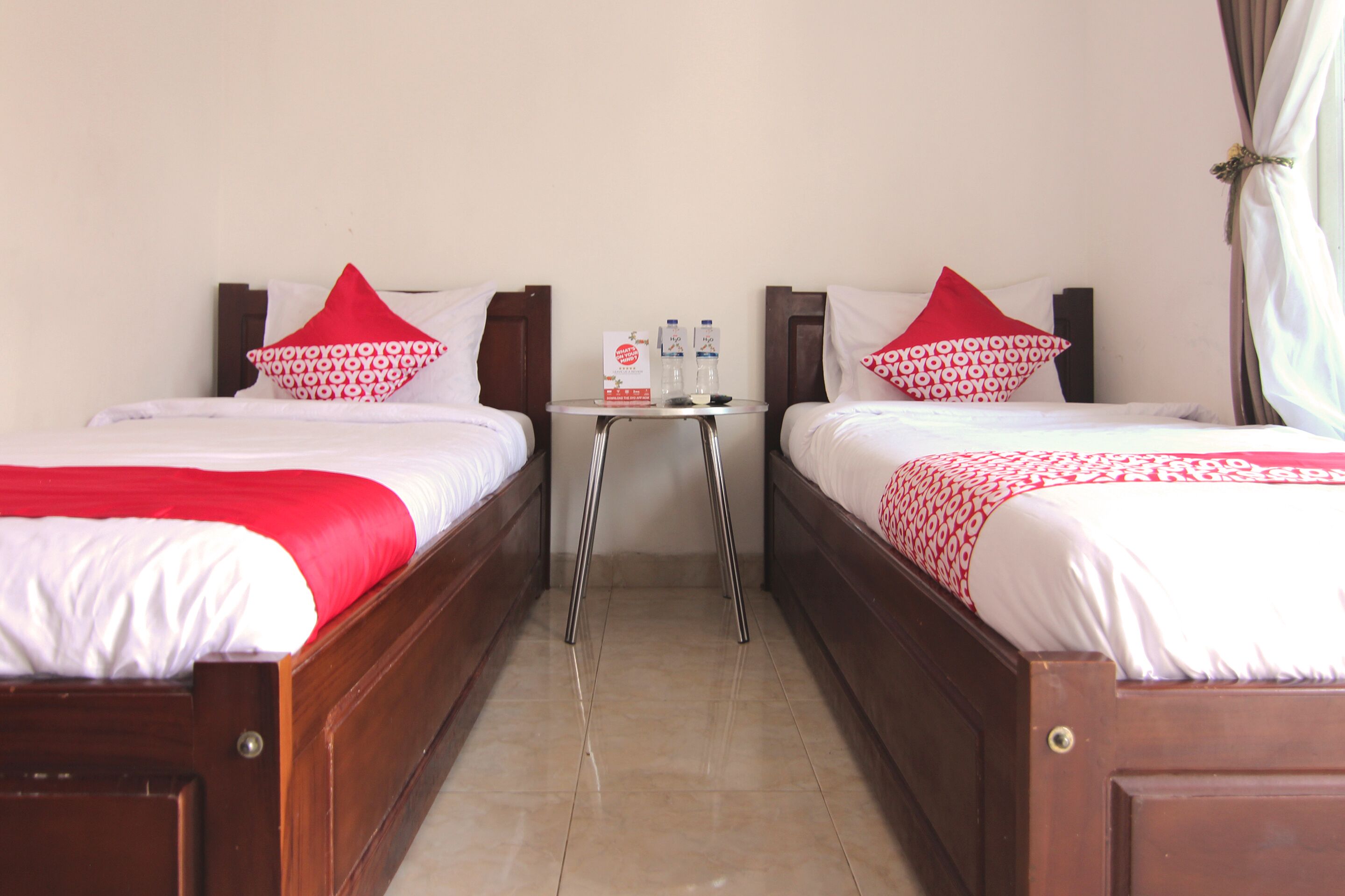 A hotel room with two beds, each with a red and white patterned pillow, a small table with a bottle and a box, and a curtain on the window.
