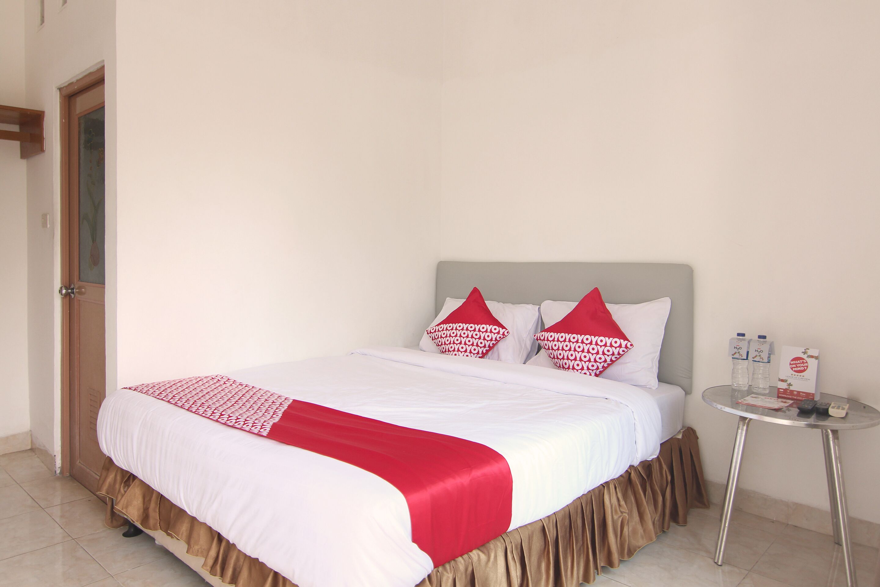 A neatly made bed with a white and red bedspread, two red and white pillows, and a brown skirt at the foot of the bed. There is a small round table with a glass top and two bottles of water and a box of tissues to the right of the bed.