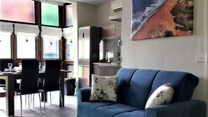 Een woonkamer met een blauwe bank, een eethoek met stoelen en een tafel, en een keuken op de achtergrond.