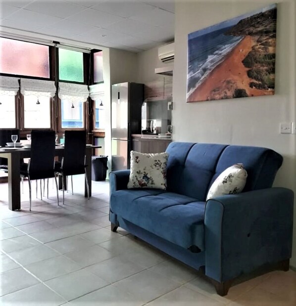 Uma sala de estar com um sofá azul, uma área de jantar com cadeiras e uma mesa, e uma cozinha ao fundo.