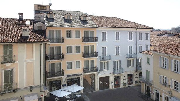 Vista de la calle con edificios de varios pisos, balcones y una cafetería con mesas al aire libre.
