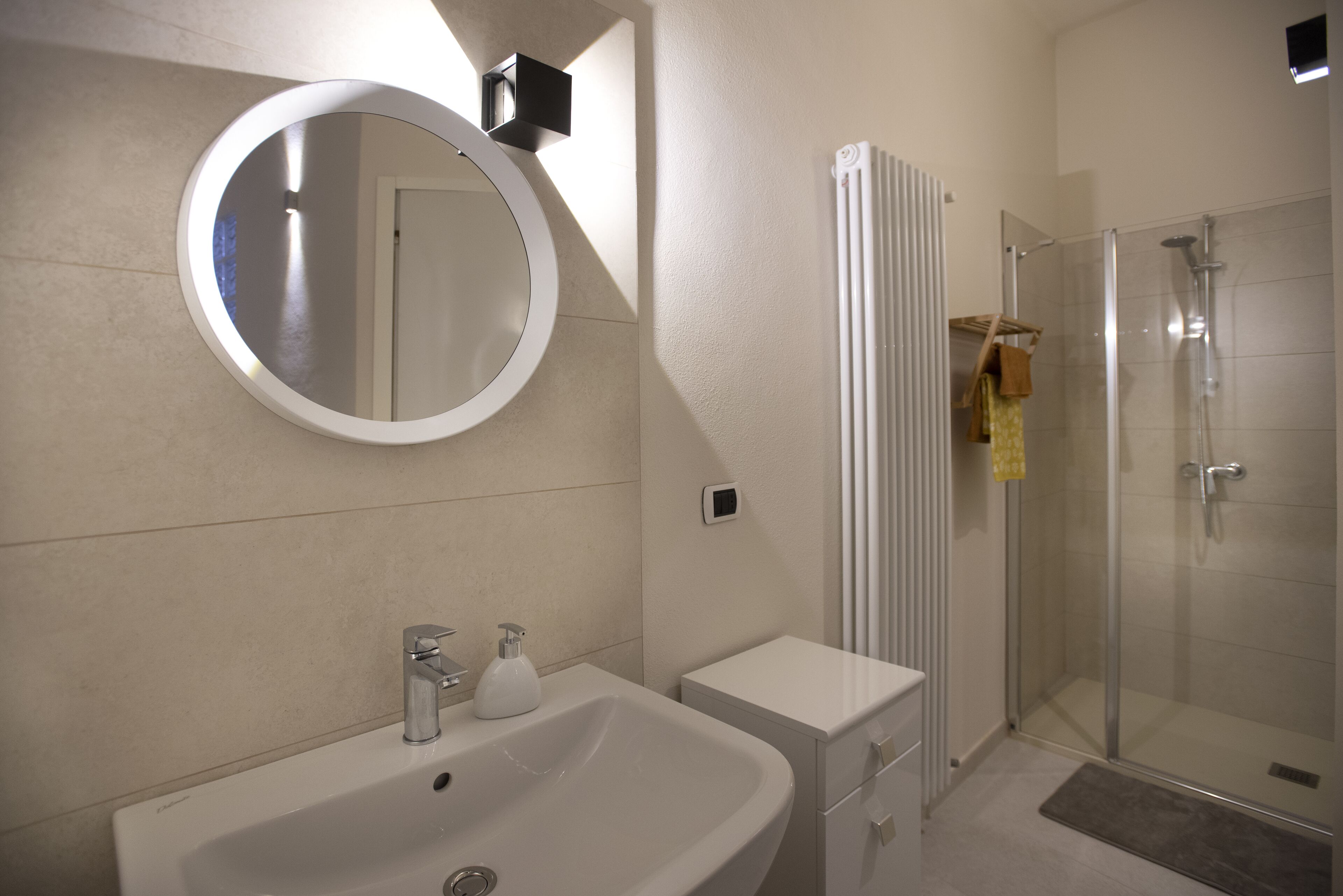 Un baño moderno con un espejo redondo, un lavamanos blanco, un mueble pequeño, una mampara de cristal para la ducha y un radiador.