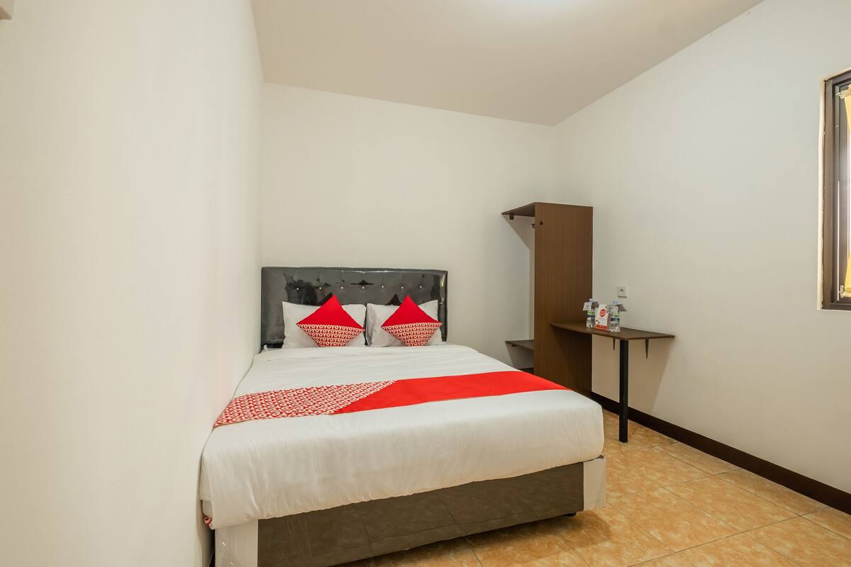 Un lit bien fait, recouvert d’une couvre-lit rouge et blanche, avec une tête de lit en bois et une petite table de chevet sur laquelle se trouve une bouteille.