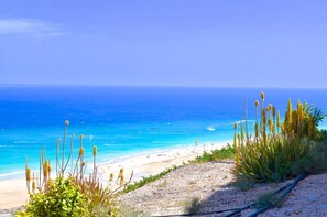 A beach with clear blue water, sandy shore, and sparse vegetation.