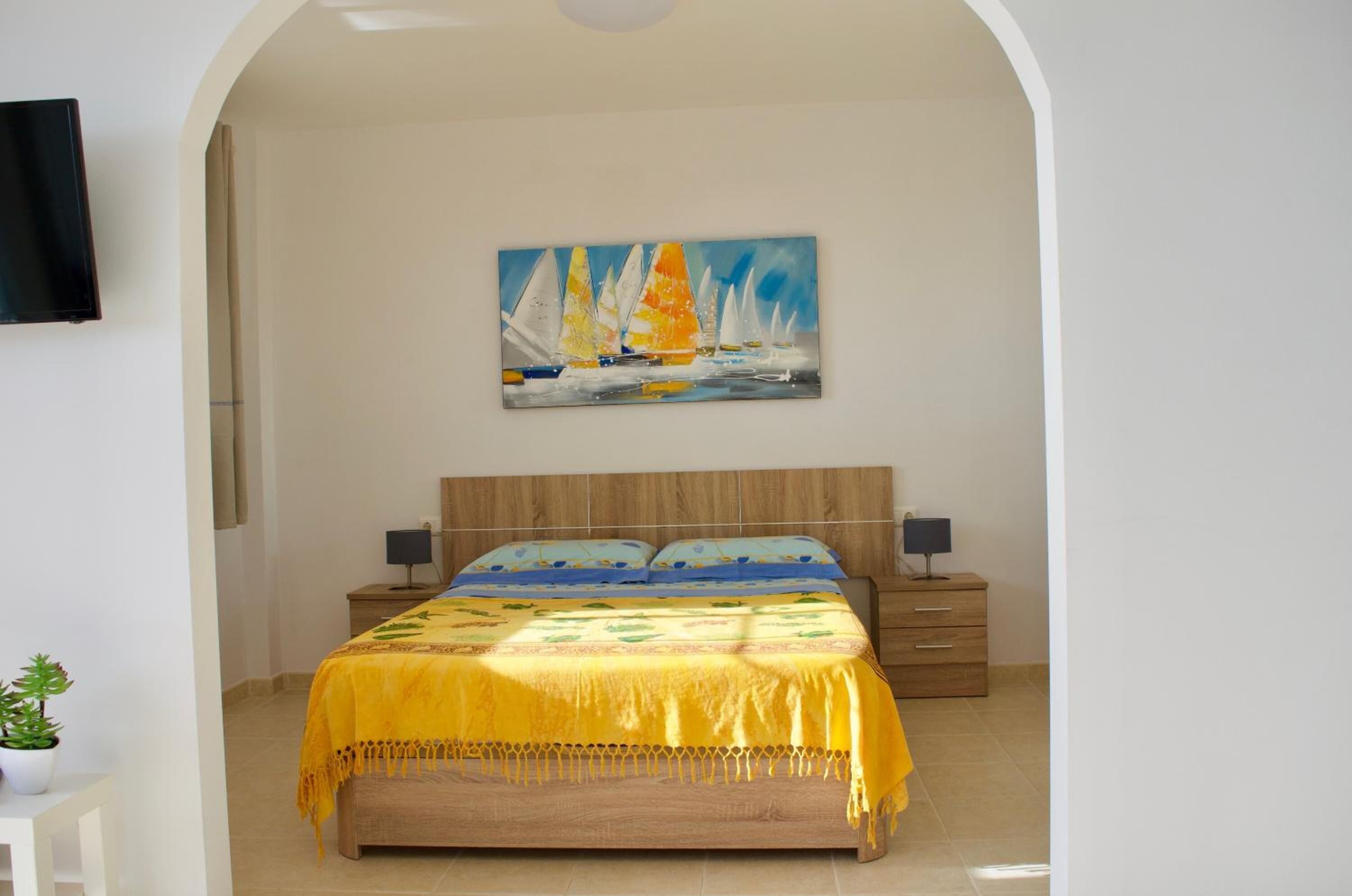 A bedroom with a wooden bed, a yellow bedspread, a painting of sailboats on the wall, and a TV mounted on the wall.