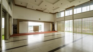 Amplo espaço interno com piso ladrilhado, um tapete vermelho central e grandes janelas que permitem entrada de luz natural.