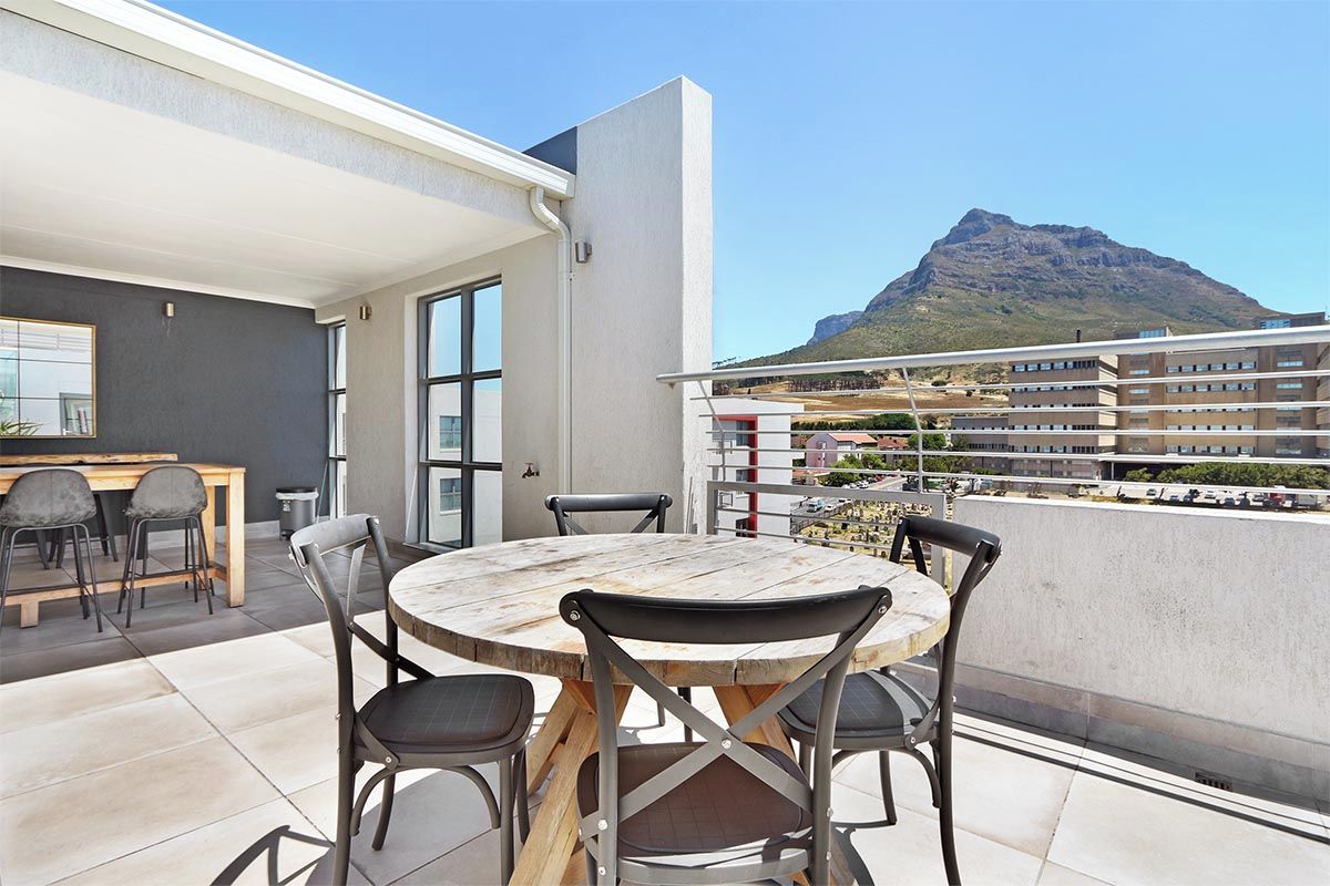 A balcony with a round wooden table and chairs, a small kitchen area, and a view of a mountain.