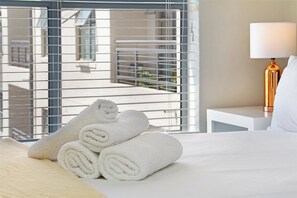 A hotel room with a bed, a window with metal blinds, a bedside lamp, and rolled towels on the bed. - The Paragon 317 (Cape Town)