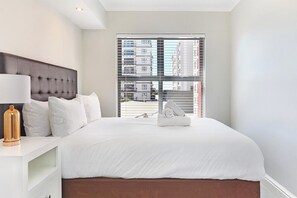 A hotel room with a large bed, a bedside table with a lamp, and a view of the city through the window. - The Paragon 317 (Cape Town)
