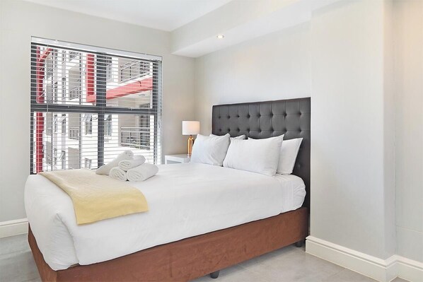 A hotel room with a large bed, a nightstand, a lamp, and a window with a view of a building. - The Paragon 317 (Cape Town)