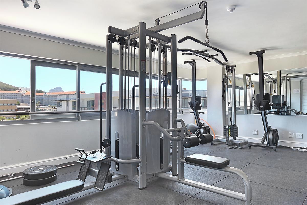 Un gimnasio con diferentes equipos de ejercicio, ventanas amplias y vistas a edificios y montañas.