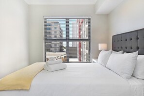 A hotel room with a large bed, a headboard, a nightstand with a lamp, and a view of buildings through a window with blinds.