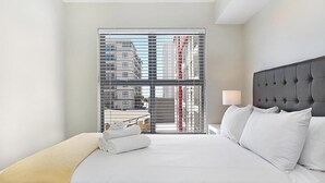 A hotel room with a large bed, a headboard, a nightstand with a lamp, and a view of buildings through a window with blinds. - The Paragon 317 (Cape Town)