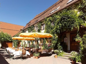An outdoor dining area with tables and chairs, covered by yellow umbrellas, and surrounded by greenery and a building with a tiled roof. - Landgasthof Adler (Rammingen)