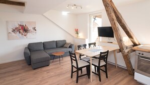 A modern living room with a gray sofa, a wooden dining table with chairs, a TV mounted on the wall, and a kitchen area with a stove. - FeWo Engler Lüneburg Wallstraße (Lueneburg)