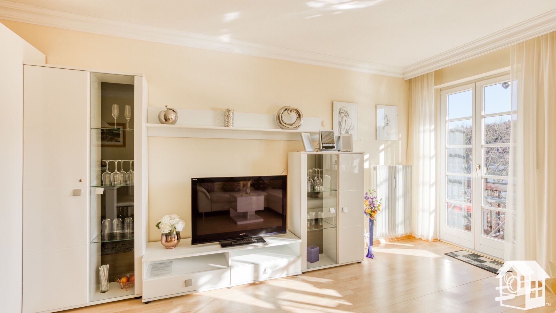 A modern living room with a flat-screen TV, glass shelving with wine glasses, and a large window with a view.