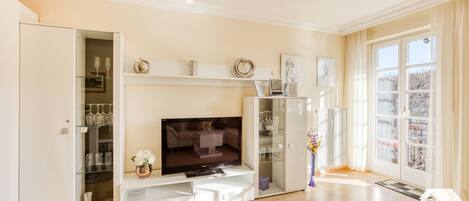 A modern living room with a flat-screen TV, glass shelving with wine glasses, and a large window with a view.