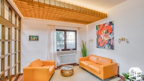 Sala de estar com dois sofás laranja, teto de madeira, uma janela grande com cortinas, um radiador e um quadro na parede.