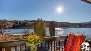 Un balcon donnant sur un lac, une balustrade en bois, une table avec un vase de fleurs et un coussin.