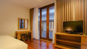 A hotel room with a bed, a TV on a wooden stand, a small table with a mirror, and a sliding glass door leading to an outdoor area. - The Chandara Villa Bali (Nusa Dua)
