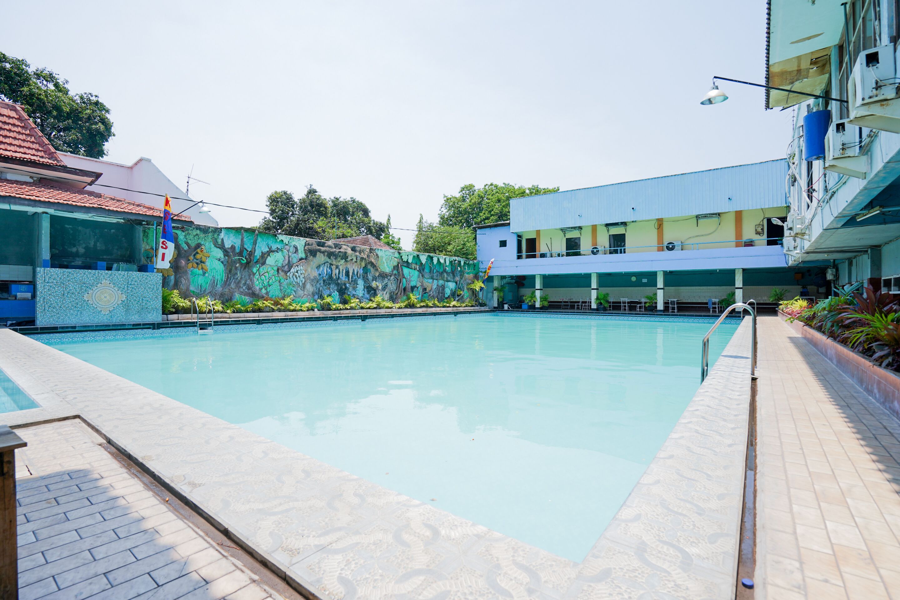 A swimming pool with a tiled deck, a mural on the wall, and a building with a blue roof in the background.