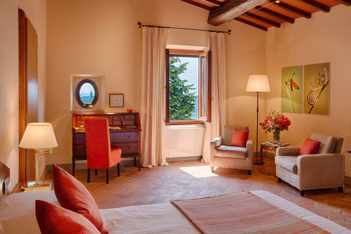 Une chambre d’hôtel comprenant un lit, deux fauteuils, un bureau avec une chaise, une fenêtre avec des rideaux, une lampe et des œuvres d’art accrochées au mur.