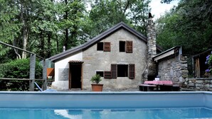 Una casa de piedra con piscina, una mesa pequeña con mantel a cuadros rojo y blanco, y una planta en maceta.