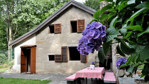 Un edificio de piedra con contraventanas de madera, un patio de piedra con una mesa y bancos, y una flor morada en primer plano.