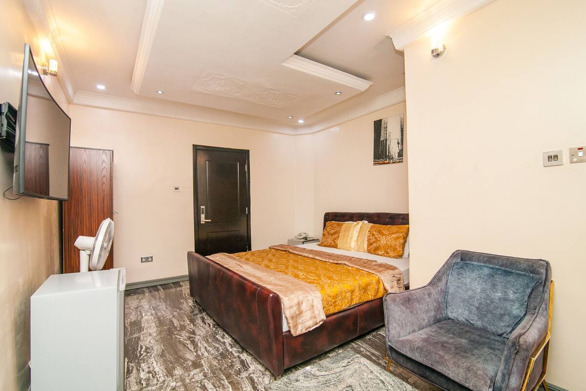A hotel room with a bed, a chair, a TV, and a fan.
